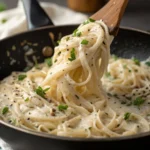 Creamy shirataki noodles being served from a pan with a wooden spoon, garnished with fresh parsley and black pepper.