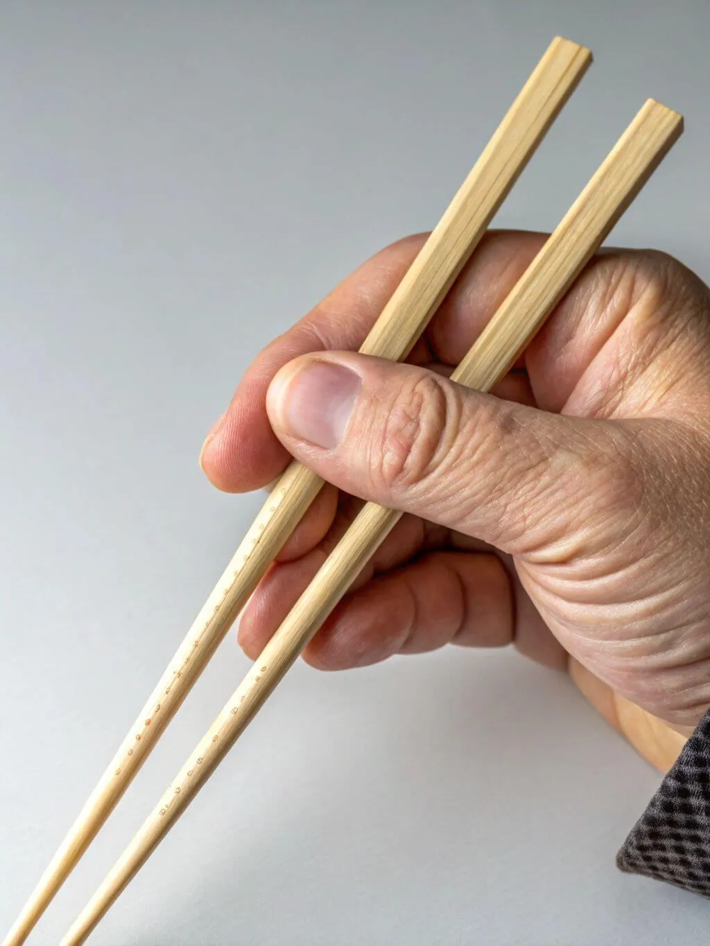 A close-up shot of a person's hand demonstrating the correct way to hold a pair of light-colored wooden chopsticks. The thumb is positioned to secure the bottom chopstick, while the index and middle fingers are poised to control the top chopstick. The background is a plain, light gray, focusing all attention on the hand and chopstick grip.