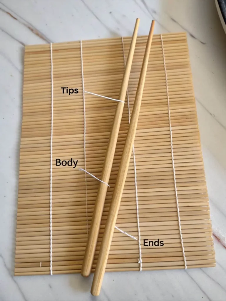 A pair of light-colored wooden chopsticks is laid flat on a bamboo sushi mat. White lines with text labels point to different sections of the chopsticks: "Tips" points to the narrower ends, "Body" points to the middle section, and "Ends" points to the thicker, blunt ends. The bamboo mat provides a textured background.