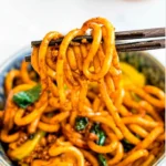 A close-up, eye-level shot of dark chopsticks lifting a generous tangle of thick, glossy, orange-brown Yaki Udon noodles from a patterned blue and white bowl. The noodles are coated in a rich sauce and mixed with visible bits of green vegetables and possibly ground meat or mushrooms. The background is a soft, bright blur, putting full emphasis on the noodles.