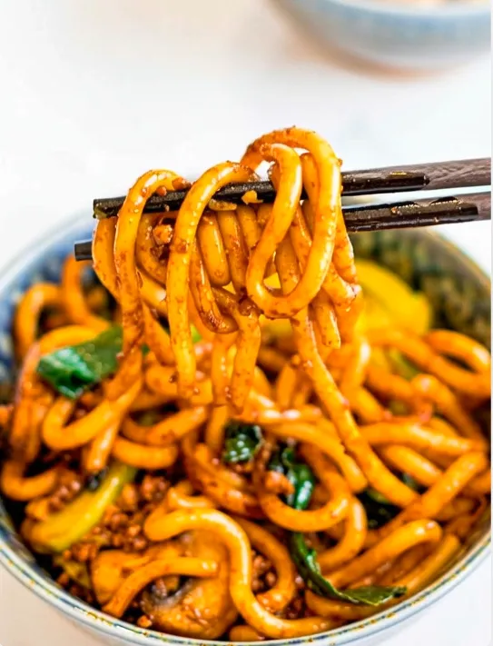 A close-up, eye-level shot of dark chopsticks lifting a generous tangle of thick, glossy, orange-brown Yaki Udon noodles from a patterned blue and white bowl. The noodles are coated in a rich sauce and mixed with visible bits of green vegetables and possibly ground meat or mushrooms. The background is a soft, bright blur, putting full emphasis on the noodles.