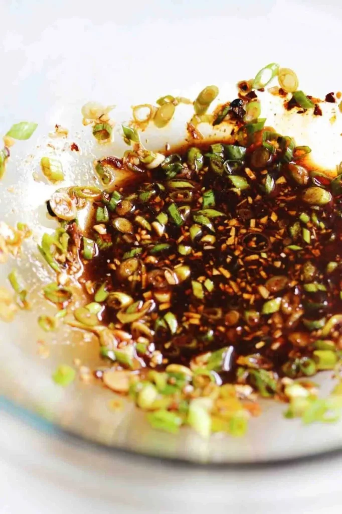 Chili oil, soy sauce, and chopped scallions combined in a glass bowl for noodle tossing