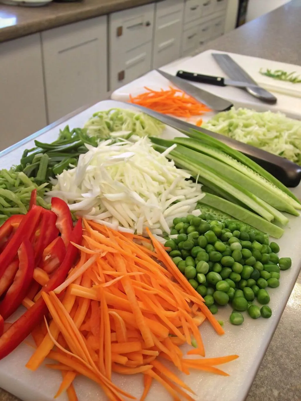 A variety of julienned vegetables, including carrots, bell peppers, green beans, cabbage, and peas, arranged on white cutting boards.