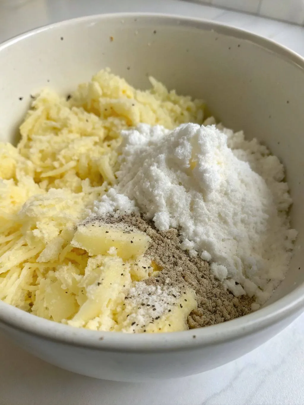 combine grated potatoes A white bowl contains a mixture of grated raw potatoes and a pile of white powder, likely flour or potato starch, with a sprinkle of dark spices.