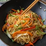 hakka noodle recipe A close-up, top-down view of cooked noodles with stir-fried vegetables being mixed in a wok with chopsticks.