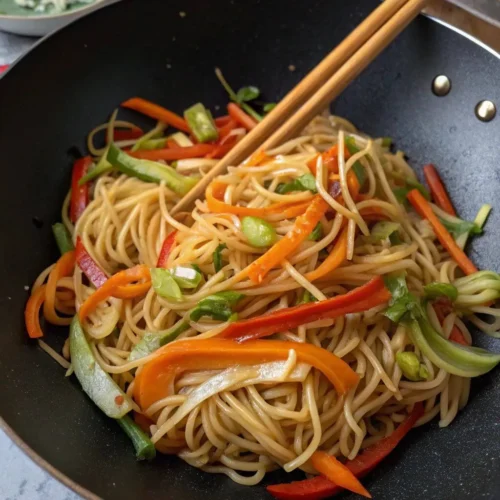 hakka noodle recipe A close-up, top-down view of cooked noodles with stir-fried vegetables being mixed in a wok with chopsticks.