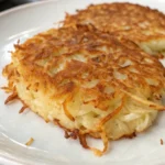 A close-up shot of a round, golden-brown hash brown patty with a perfect, crispy texture, resting on a beige plate. A second, blurry hash brown is visible in the background.