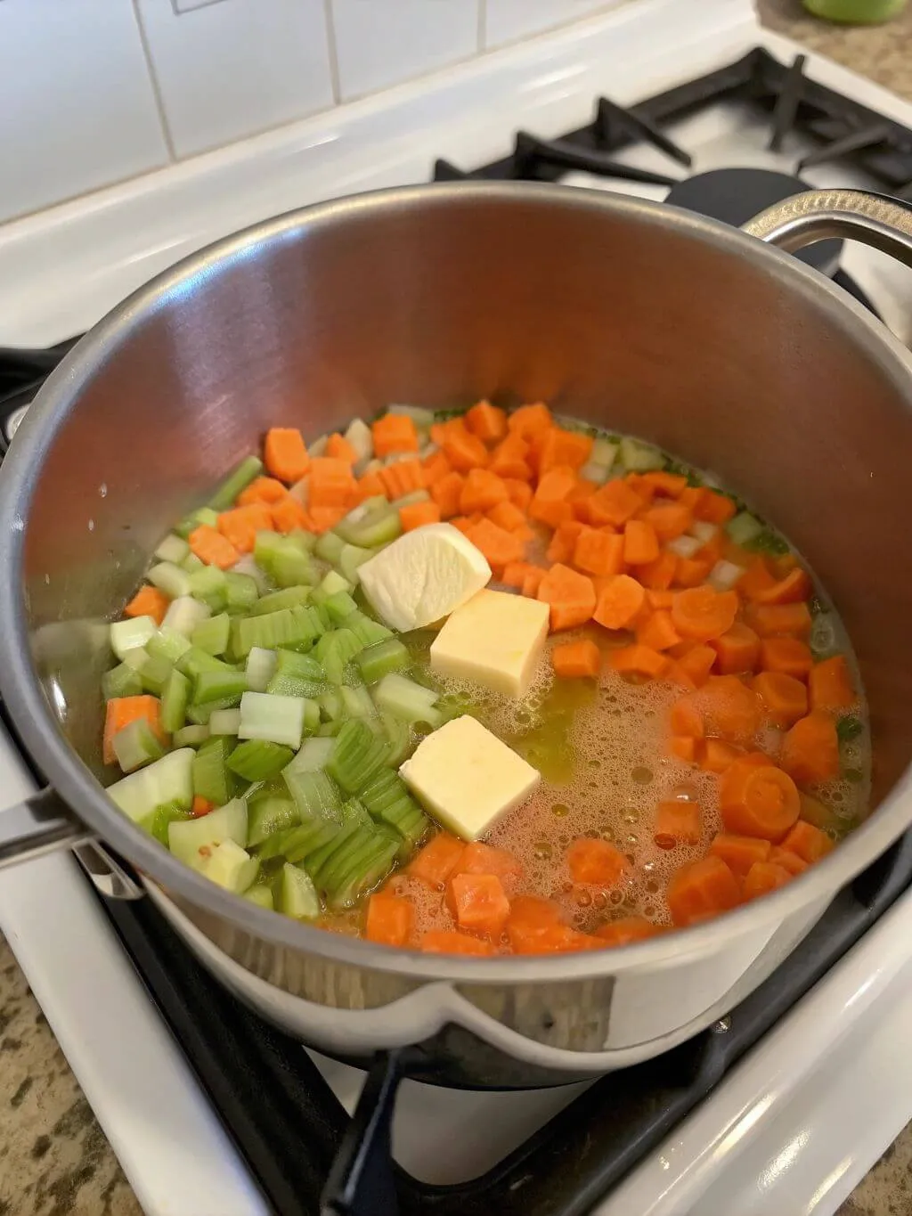 Sauté the Vegetables A close-up shot of chopped carrots and celery being sautéed in a stainless steel pot with two pats of butter. The vegetables are glistening and a few bubbles show that the pot is on a stove burner